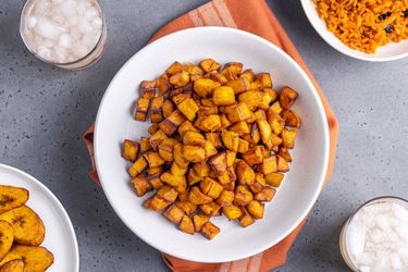 A bowl of fried dodo in a white bowl on top of an orange towel, surrounded by a bowl of jollof and a second plate of plantains.