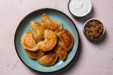 Serving plate of pan-fried pierogi with ramekins of sour cream and cooked onion on the side.