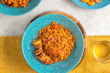 Overhead view of Jollof rice on a bright blue plate on top of a yellow table runner next to a glass of water