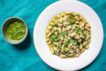 顶视图的大蒜柱身香蒜沙司in a small bowl next to a wide rimmed, shallow bowl of cooked white beans and pesto