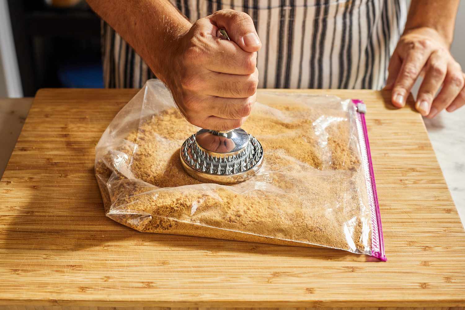 一只手正在用一个银色的短柄肉捣碎器捣碎塑料袋里的全麦饼干屑