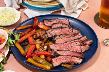 Platter of grilled skirt steak fajitas, peppers, and onions, with tortillas, cheese, and other toppings in the background.