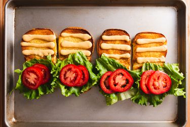 四个烤部rger buns open on a baking sheet, each one with three lines of homemade Shack Sauce on the top half of the bun, and lettuce and tomato on the bottom half of the bun.