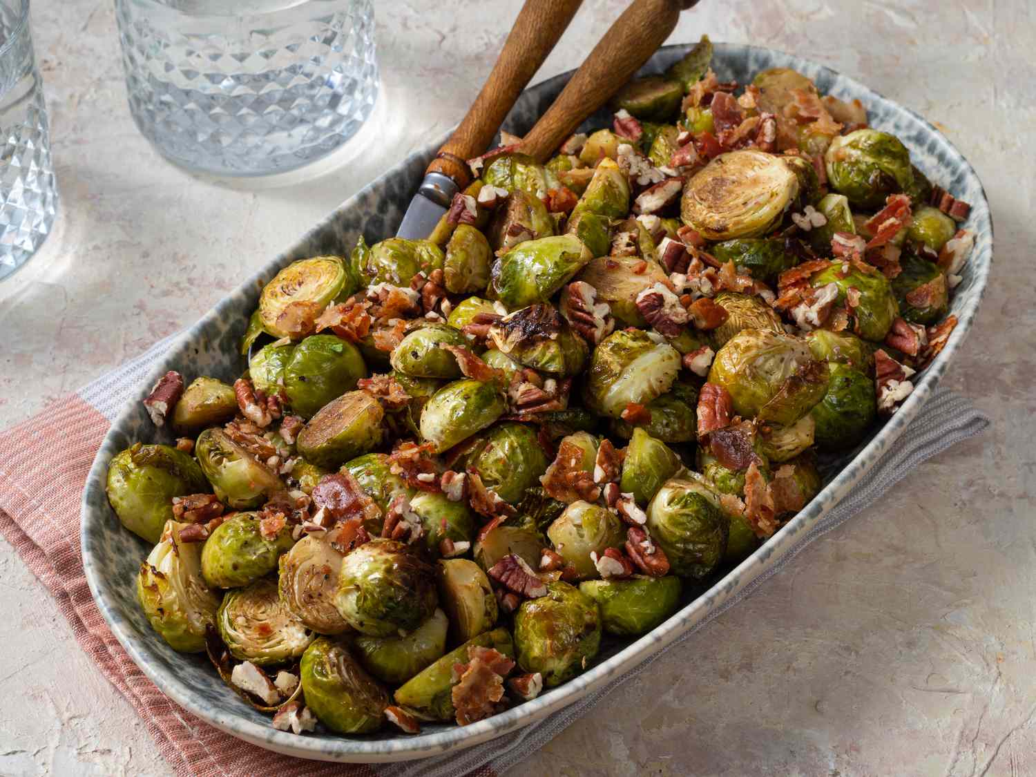 Brussels sprouts in a ceramic serving dish with tongs, a napkin, and glass cups on the periphery