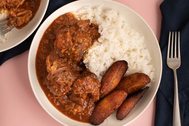 Rabo encendido with white rice and maduros served in a white bowl on a pink countertop.