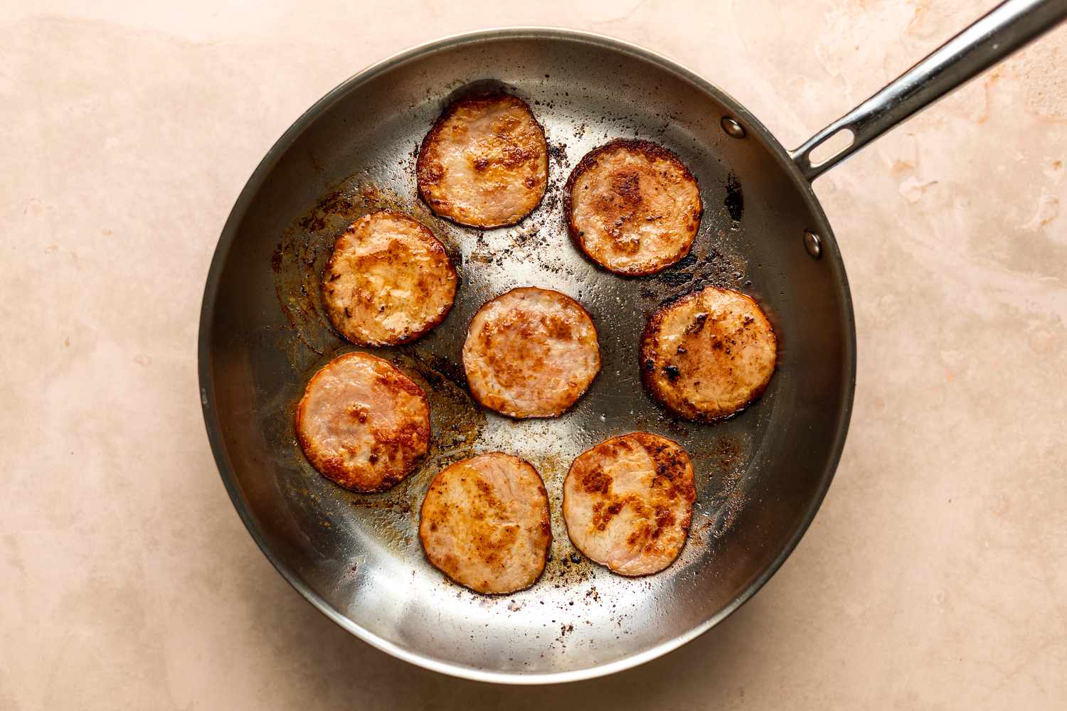 A stainless steel skillet holding eight slices of Canadian bacon which has been browned.