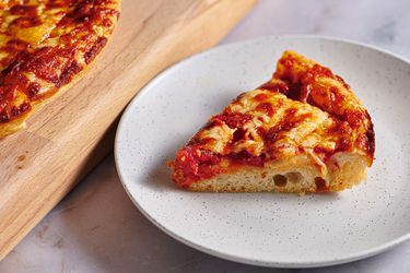 A slice of New England Greek-Style pizza on a speckled ceramic plate. There is a cutting board holding more pizza on the left hand side of the image