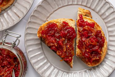 Two slices of bread topped with tomato and onion jam on a ridged cream colored ceramic plate. In the left corner of the image is a glass jar filled with more jam, and in the top left corner is a portion of another plate with more toast on it.