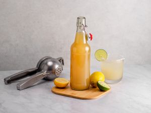 A bottle of sour mix on a wooden cutting board with a lemon, sliced lime, citrus squeezer, and a mixed drink in a glass.