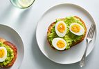 Halved hard-boiled eggs topped with flaky salt and placed on bread topped with mashed avocado. The food is placed on a white ceramic plate with a metal fork.