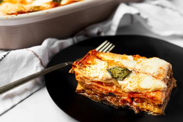 Slice of lasagna served on a small plate, with casserole dish holding lasagna peeking into the frame