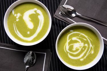 Overhead view of garlic scape soup in two white bowls on a black with thin white stripe background and two grey napkins and spoons