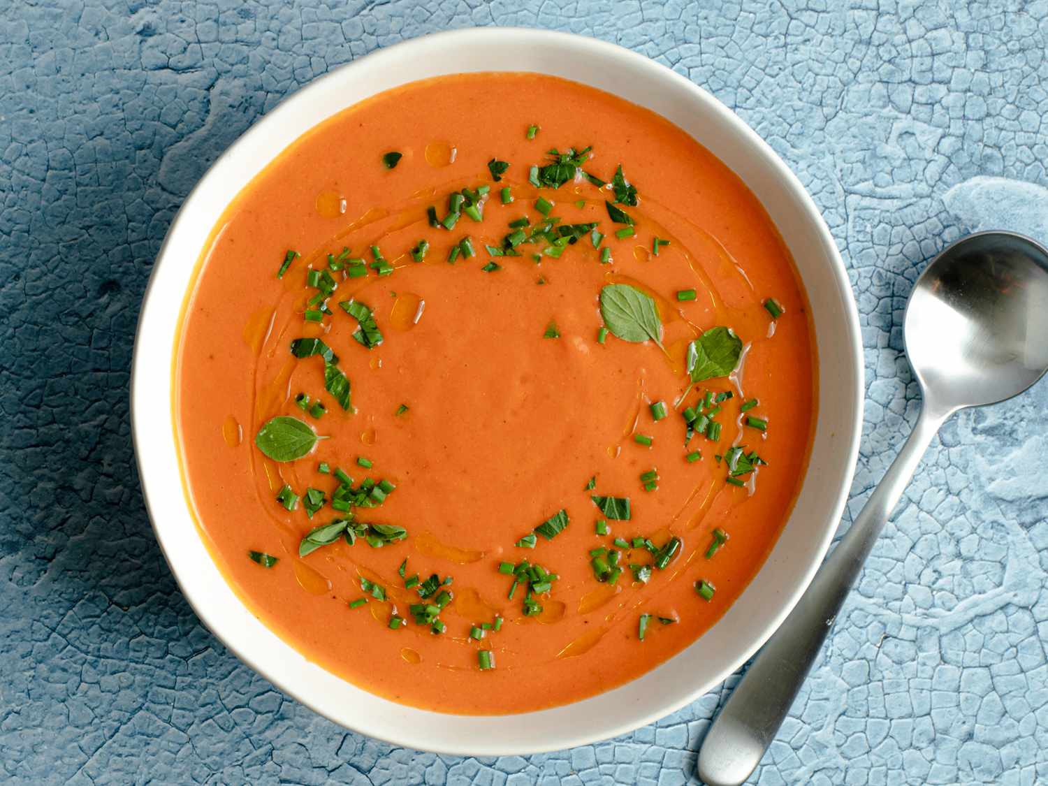 A bowl of gazpacho sprinkled with chopped herbs. The bowl is on a mottled blue surface and there is a metal spoon to the right of the bowl.