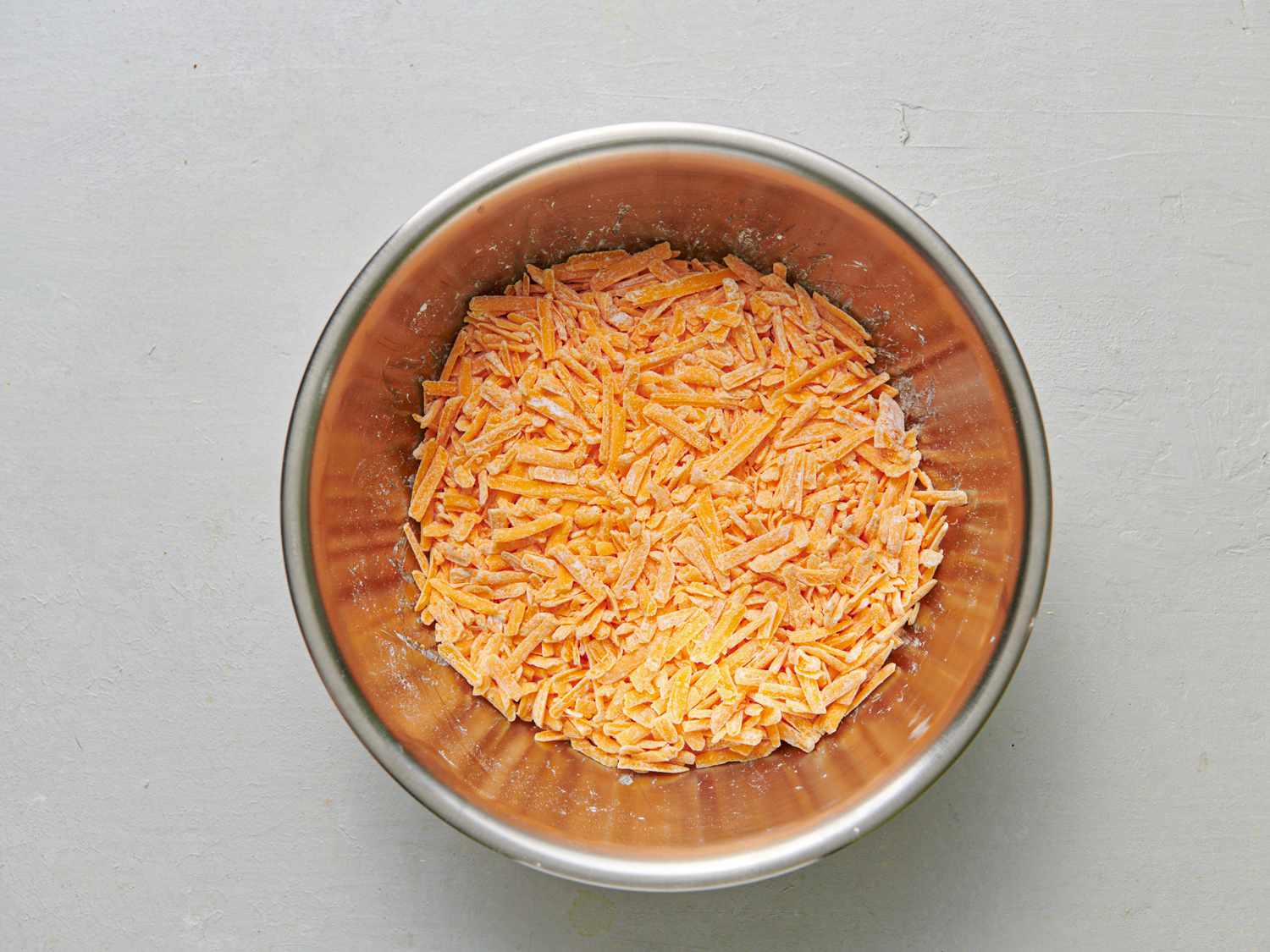 A stainless steel bowl holding shredded cheese tossed with corn starch.