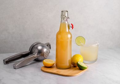 A bottle of sour mix on a wooden cutting board with a lemon, sliced lime, citrus squeezer, and a mixed drink in a glass.