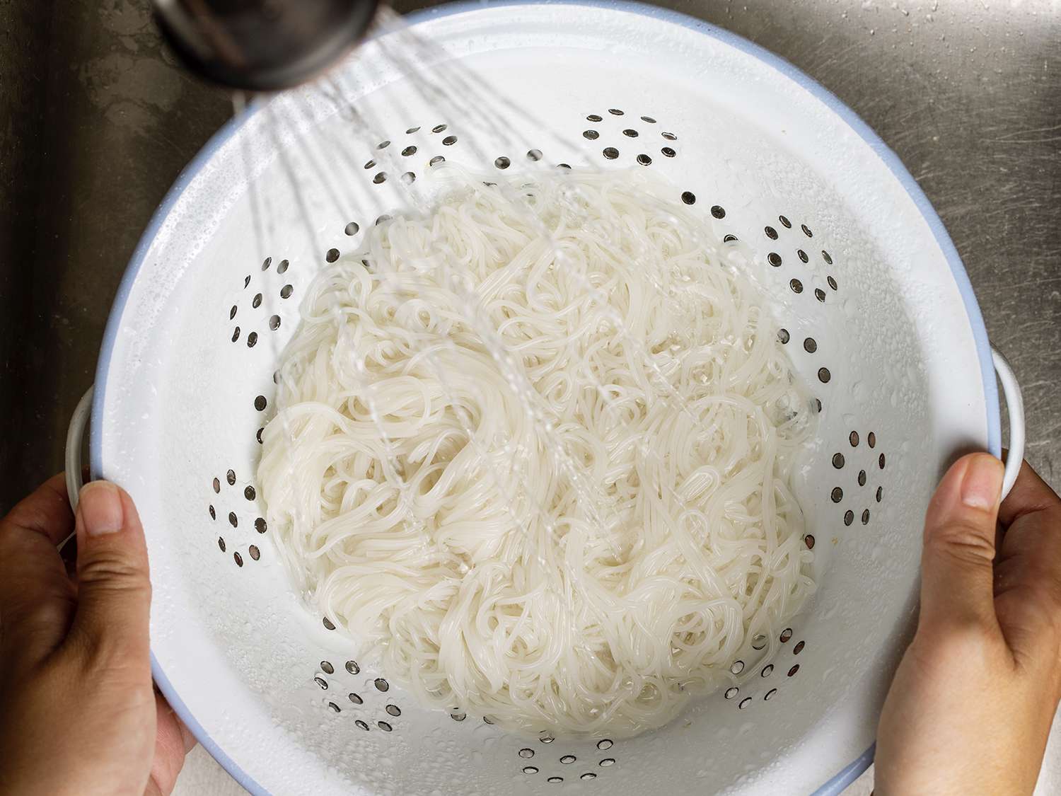 Rice vermicelli being rinsed under water in a colander.