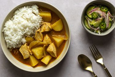 A bowl of Thai chicken yellow curry in a cream colored ceramic bowl. On the right side of the image is a smaller bowl holding the accompanying cucumber salad. There are a fork and spoon in the bottom right hand corner of the image.