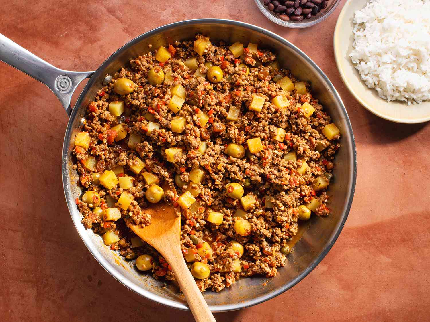 A stainless steel pan containing the cooked picadillo, with bowls holding white rice and black beans on the periphery.