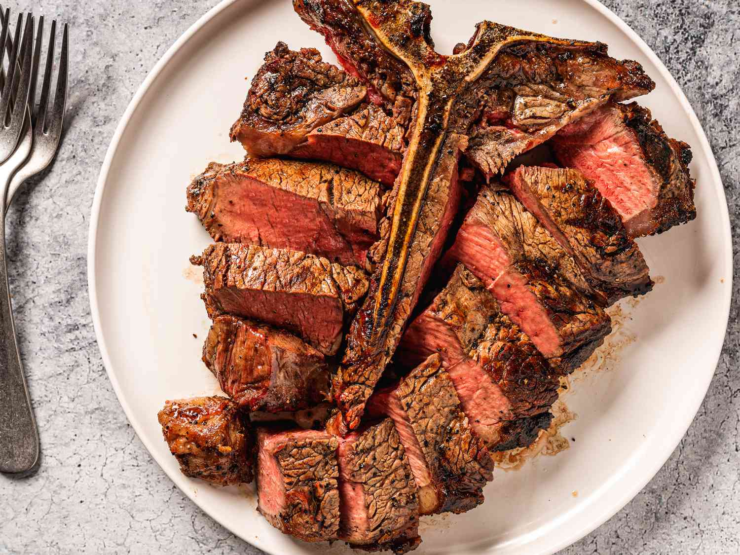 A t-bone (porterhouse) steak on a white plate, with the sliced meat placed around the bone. On the lefthand side of the image are several metal forks stacked on top of each other.