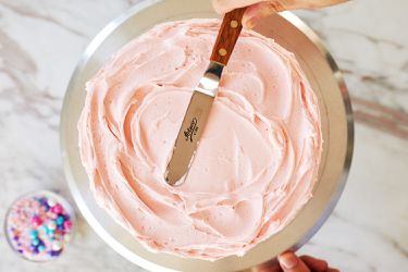 A person using an offset spatula to frost a cake on a cake stand