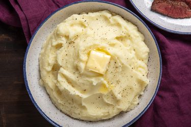 A bowl of fluffy mashed potatoes sits on a table. It's topped with a pat of melting butter and a grinding of fresh black pepper.