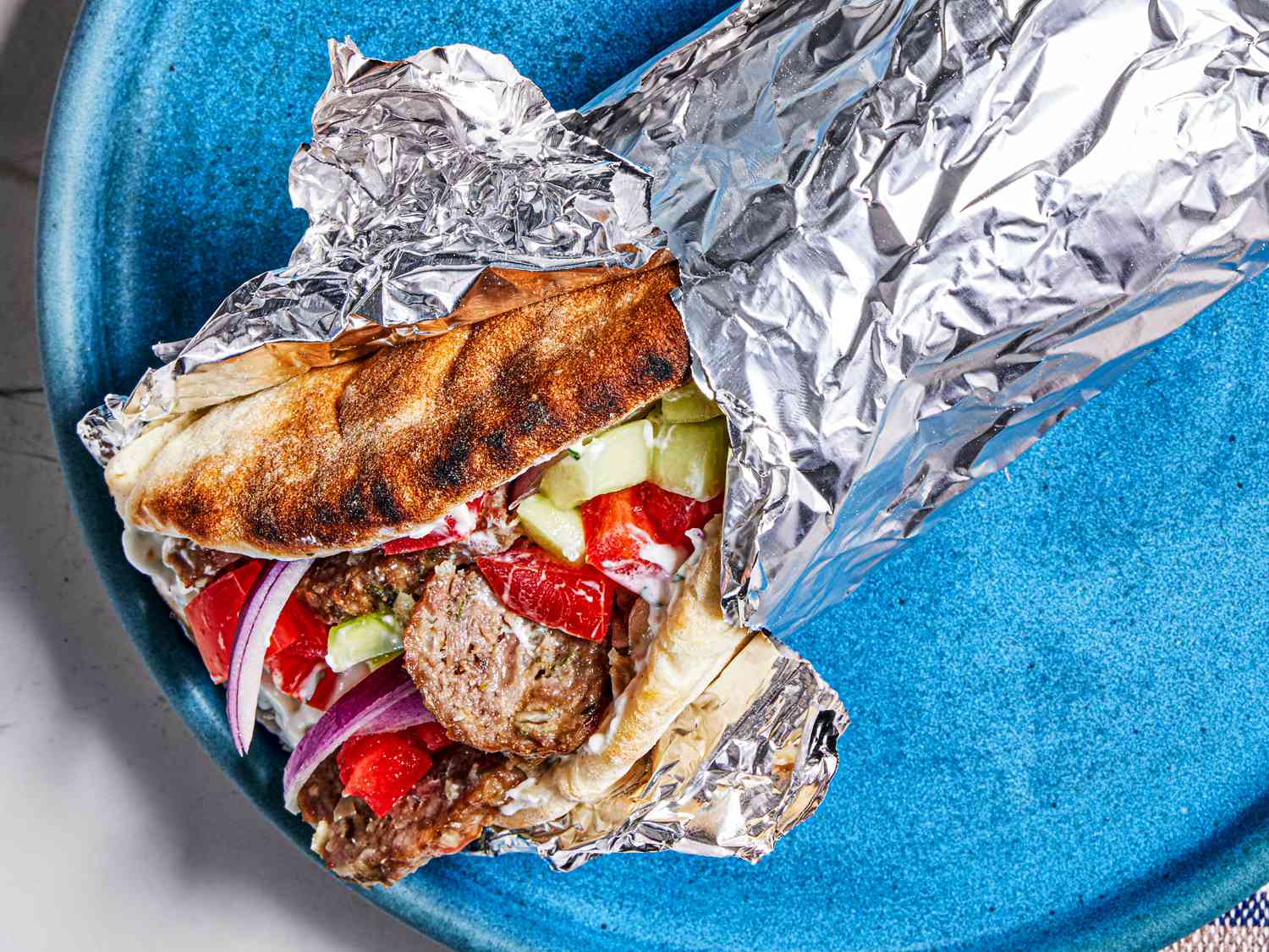 Greek-American Gryo wrapped in aluminum foil on a bright blue plate.