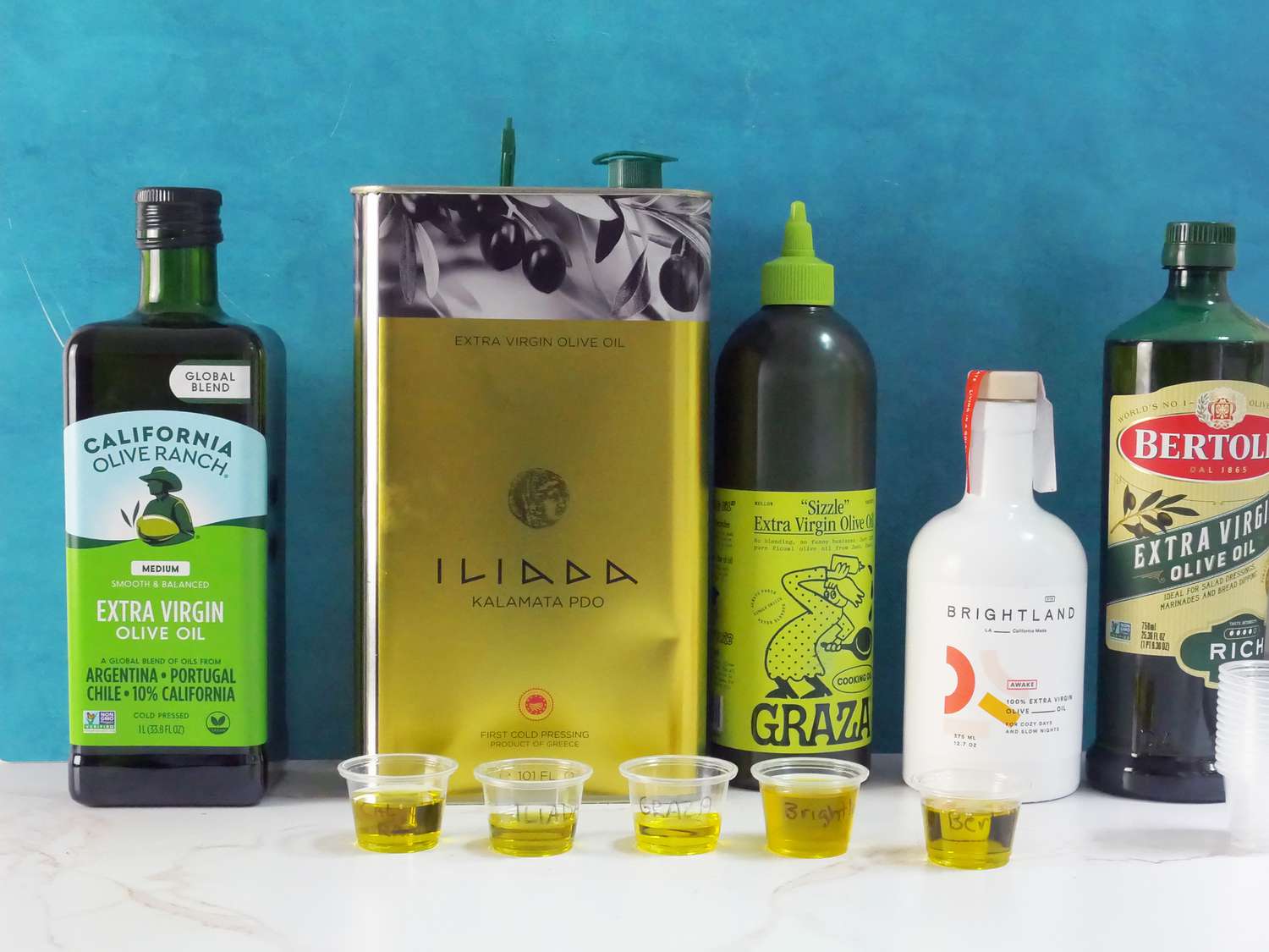 five olive oils against a blue backdrop with small tasting cups of olive oil in front