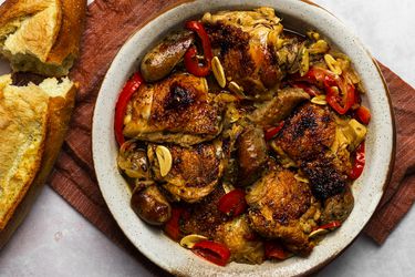 Chicken scarpariello in a round ceramic platter. The platter is on a maroon colored piece of cloth, and there is a broken loaf of bread on the left hand side of the image.