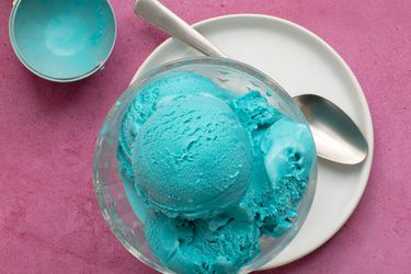 Blue moon ice cream in a glass bowl, set on a white ceramic plate which also holds a metal spoon. The background is pink and in the top left of the image is the ice cream scoop which had been used to scoop the ice cream.