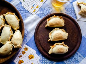 Overhead view of a table scene. Two plates of empanadas are placed on top of a traditional Argentinian fabric with a blue and white checkered. In the middle of the frame is a dark wooden table with 3 empanadas in a line, surrounded by a beer, playing cards and a half eaten empanada.