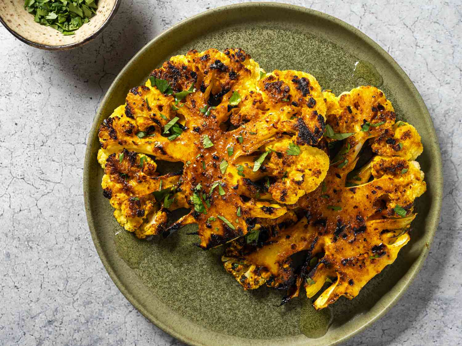 A head of spiced and grilled cauliflower, cut in half lengthwise and overlapping on a green ceramic plate. At the top left edge of the image is a small ceramic bowl holding chopped herbs, which are also sprinkled across the surface of the cauliflower.