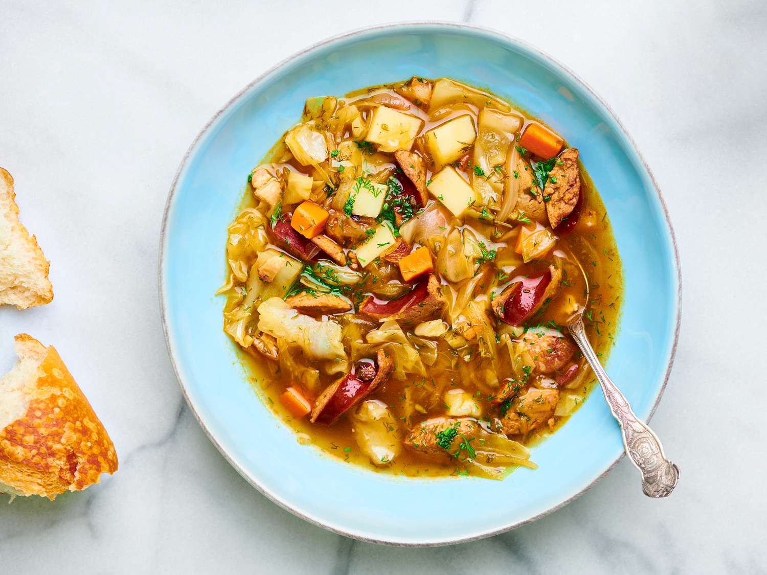 Polish potato, kielbasa, and cabbage soup in a blue bowl, with torn bread on the left periphery.