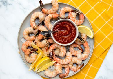 Shrimp cocktail on a ceramic plate with cocktail sauce and sliced lemons, on a stone background with a yellow dish towel.