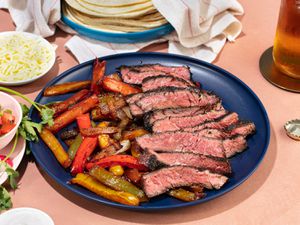 Platter of grilled skirt steak fajitas, peppers, and onions, with tortillas, cheese, and other toppings in the background.