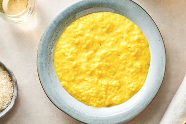 A round ceramic plate holding creamy saffron risotto. There is a glass of liquid in the top left corner and a small bowl of grated cheese on the bottom left edge of the frame.