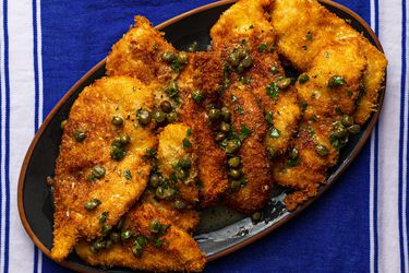 一个椭圆盘topp的炸鸡片ed with lemon-butter pan sauce containing capers. The platter is on a blue and white dish towel.