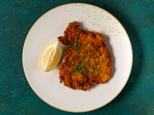 A golden brown fried chicken schnitzel cutlet with a lemon wedge on a gold and white ceramic plate on a teal-colored textured background.