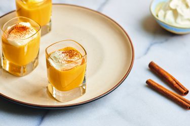 Three small glasses filled with DIY Advocaat and topped with whipped cream and dusted with cinnamon, on a ceramic plate. On the right periphery are a couple of sticks of cinnamon and a small bowl of whipped cream.