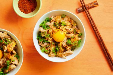 Oyakodon, in a white ceramic bowl alongside wooden chopsticks, a small ceramic bowl holding togarashi seasoning, and an additional bowl of oyakodon off to the left side.