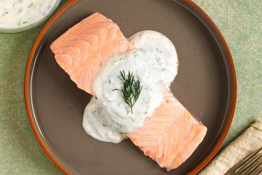 Poached salmon topped with dill-yogurt sauce on a dark plate with a red rim. There is a fork sitting on a napkin in the bottom right corner, and a small bowl of additional sauce in the top left corner.