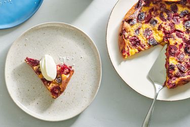 A slice of cherry clafoutis on a speckled ceramic plate on the left side of the image. On the right side is the remaining clafoutis, showing a slice has been removed.