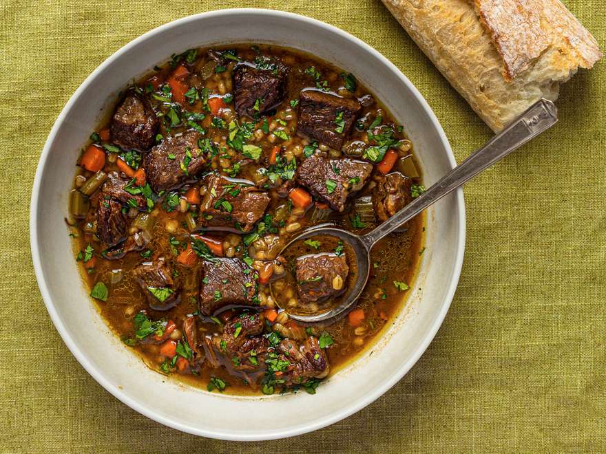 A white porcelain bowl holding beef barley soup. There's a metal spoon in the bowl and the bowl is palced on a green cloth. There is also part of a baguette in the top right corner of the image.