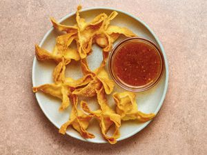 Golden, fried crab rangoons on a blue ceramic plate which also holds a small glass bowl of sauce, on a pink stone background.