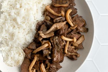 A closeup shot of stir-fried beef with mushrooms and butter. The beef and mushrooms are plated in a round white bowl with white rice on a white tile background, and the shot shows how the mushrooms have been seared, while the feed is tender and in a somewhat glossy sauce.